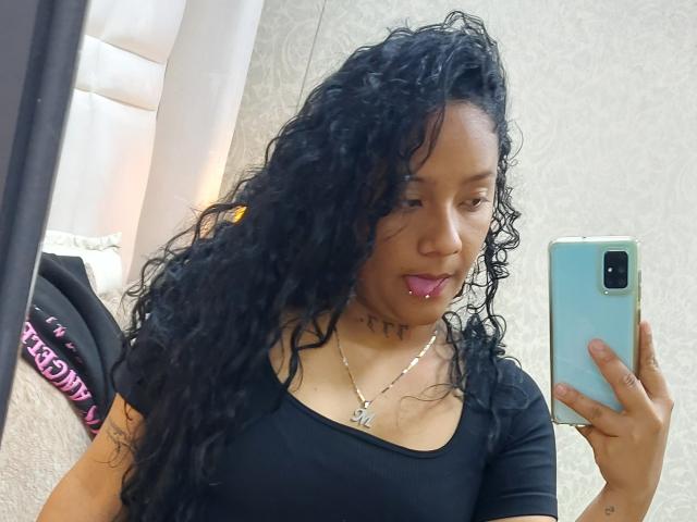 Kimbherly, avec ses cheveux noirs bouclés, son rouge à lèvres rose, son haut noir, son collier, tenant un téléphone, regardant dans le miroir, montrant de petites seins, peau brun clair, tatouage sur le bras, et rouge à lèvres rose..