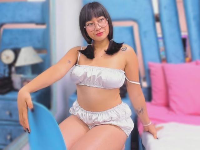 Genesiss, avec ses couettes noires, ses lunettes et sa lingerie blanche à volants, montrant ses seins et ses fesses, assise sur une chaise bleue, avec des tatouages sur son bras, dans une chambre colorée..