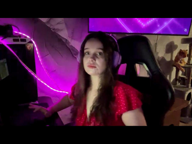 ReneBoehringer with long brown hair, wearing a red shirt, sitting in a black chair, using a computer, with pink neon lights and a headset on.