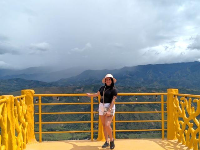 Catalina Rivero está em frente a um corrimão amarelo, vestindo uma blusa preta, shorts brancos, sapatos pretos e um chapéu branco, com uma paisagem de montanhas nubladas ao fundo. Ela está olhando para a câmera..