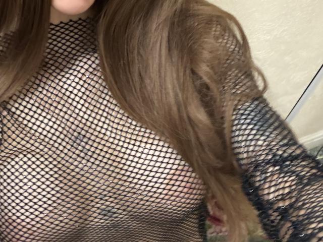 SensualMila wearing a black fishnet top, showing her tits through the net. Long brown hair covering one side. Soft light, slightly blurred. SensualMila's nipples are barely visible.