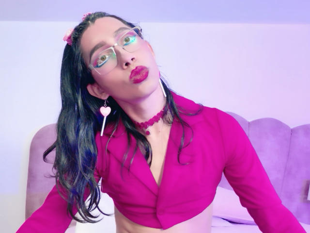 AlexaMissSx, con labios rosados y voluminosos, vistiendo una camiseta rosa corta, un collar rosa, aretes rosas y pinzas para el cabello rosas, mirando hacia arriba con ojos verdes y gafas, sentada en un sofá morado..