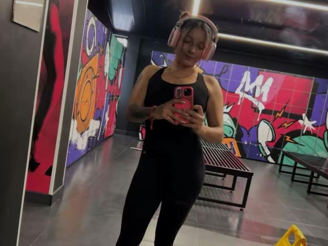 SaarahJones, with headphones on, wearing a black sports bra and leggings, taking a mirror selfie in a gym, showing off her toned arms and curvy ass.