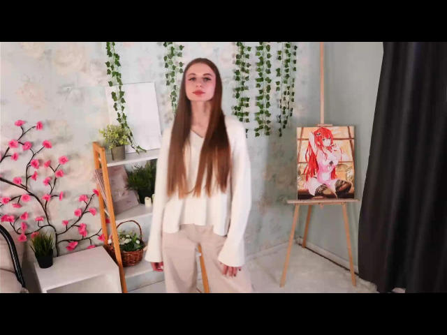 LovinValentine with long brown hair, white blouse, and beige pants stands in front of an easel with a red-haired painting. Pink cherry blossoms and green vines are visible.