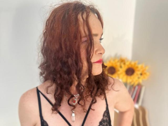 MishellBons met krullend bruin haar, gekleed in zwart kant lingerie, met rode lippenstift en een ketting, kijkt opzij, met zonnebloemen op de onscherpe achtergrond..