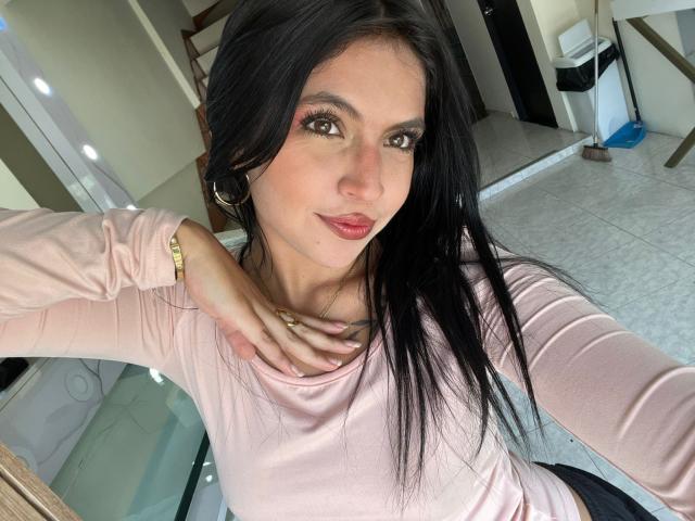 NyraVanel con lunghi capelli neri, una maglietta a maniche lunghe rosa, un braccialetto dorato, orecchini a cerchio, rossetto rosa, occhi marroni, una mano sul petto, sorridente, in interni, selfie..