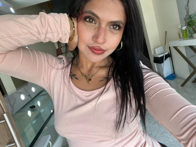 NyraVanel con lunghi capelli neri, una maglietta a maniche lunghe rosa, orecchini a cerchio dorati, una collana, tatuaggi, rossetto rosso, braccio alzato, sorridente, occhi marroni, seno di media grandezza, pelle chiara, selfie scattato in interni..