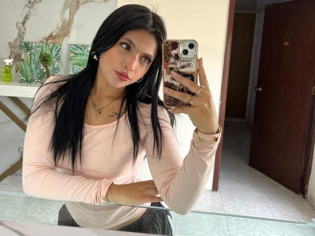NyraVanel, con lunghi capelli neri, indossa una maglia a maniche lunghe rosa, tiene il telefono davanti allo specchio, mostrando il suo seno piccolo, tiene il telefono con una custodia a pois di leopardo, ed è in piedi nel bagno..