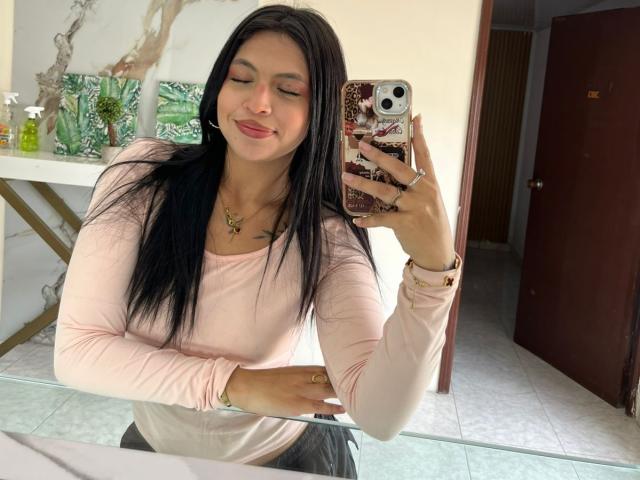 NyraVanel, com cabelos longos e pretos, blusa rosa de manga comprida e batom vermelho, sorrindo em uma selfie no espelho, segurando o celular com uma capa brilhante, exibindo seios pequenos e um traseiro atraente..