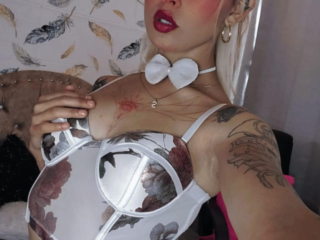 LunaLindaHot, con tatuaggi, indossa un reggiseno bianco con fiori, un collare con fiocco e rossetto rosso, tiene un seno con la mano, mostrando un tatuaggio sul petto..