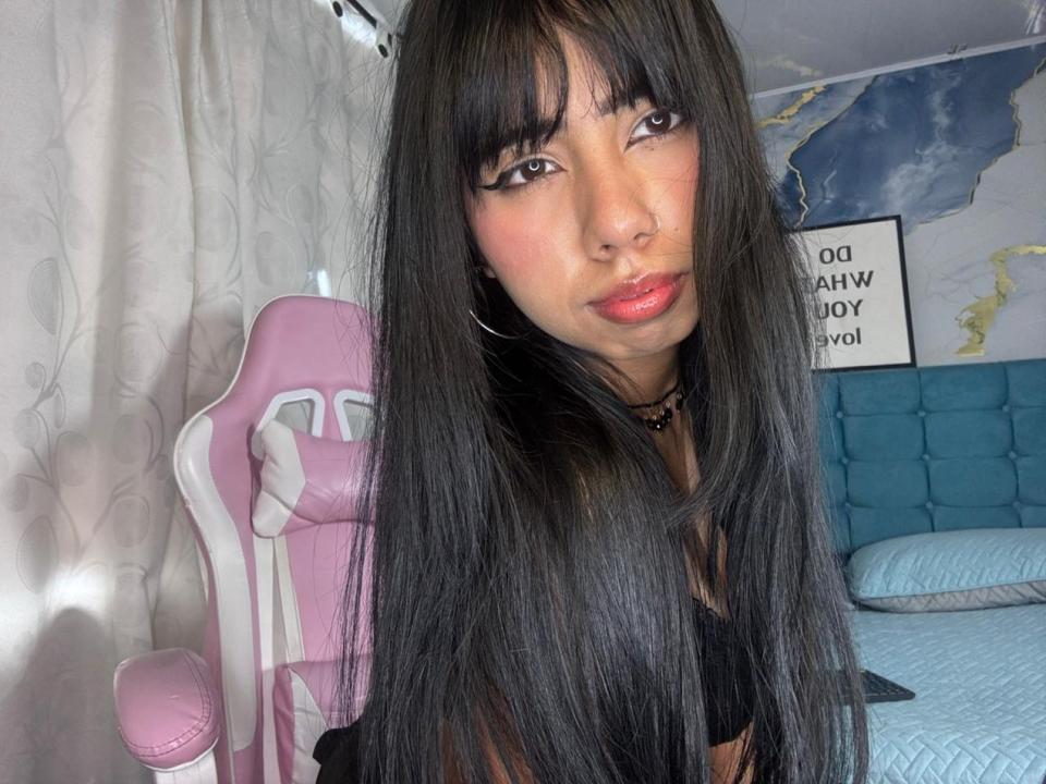 AyameFoxy mit langen schwarzen Haaren, pinkem Lippenstift und Ohrringen, sitzt auf einem pinken und weißen Gaming-Stuhl und zeigt ihr schmollendes Gesicht und ihr schwarzes Oberteil. Schlafzimmer-Szene..