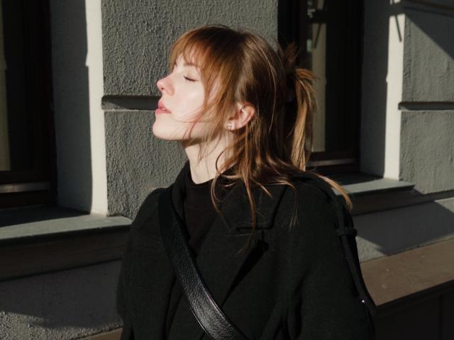 Eleonor Yoneoka, met gesloten ogen, draagt een zwarte jas, haar roze lippen zijn lichtjes open, en haar bruine haar zit in een paardenstaart. Een zwarte band kruist haar borst. Zonlicht schijnt op haar gezicht..