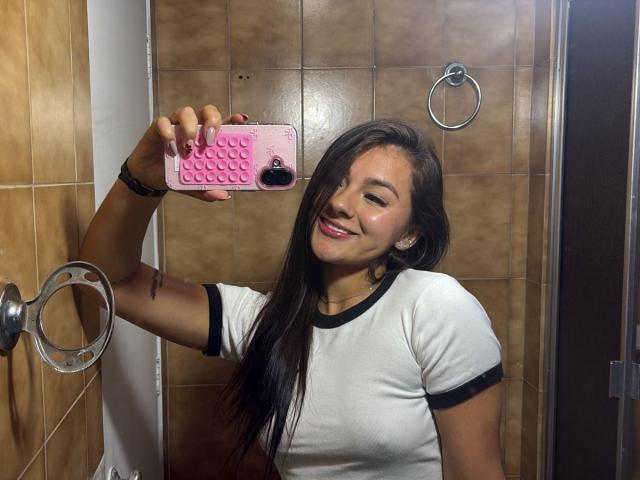 AlessiaFerry, con lunghi capelli neri, sorridente, sta scattando un selfie allo specchio con un telefono rosa, indossa una camicia bianca con bordi neri, mostrando i suoi piccoli seni e la sua pelle marrone..