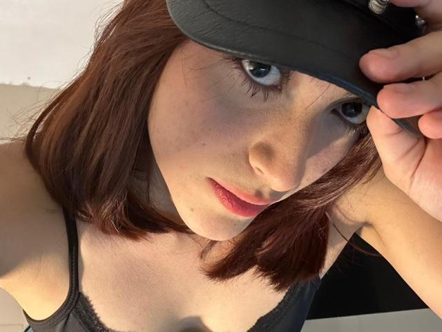 SpencerLe, con cabello castaño, ojos azules y labios rosados, vestida con una camiseta negra y una gorra negra, mirando hacia arriba con un ligero ceño fruncido, sosteniendo la gorra con la mano derecha..