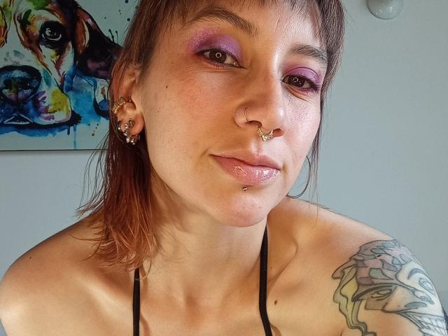 AliRousse tiene sombra de ojos morada, un piercing en la nariz, un piercing en el labio y un tatuaje en el hombro derecho. Lleva un top de tirantes negros y tiene el pelo castaño..