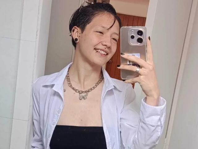 LissyLuxe with short black hair, smiling, wearing a white shirt over black top, silver necklace, black nail polish, holding phone in mirror selfie, showing her tits.