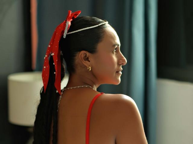 SolCozzy con i capelli scuri, un fiocco rosso a pois, un reggiseno rosso e una fascia per capelli con catena, che mostra la schiena e il fianco, piccoli orecchini dorati, che guarda a destra..