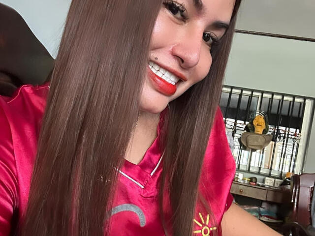 FeminaConnyX with long brown hair, red lipstick, nose piercing, smiling, wearing pink shirt, cute, sexy, showing teeth, upper body, indoors, soft lighting, casual selfie, confident.