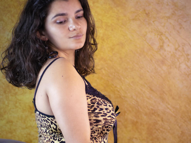 BulmaHot with wavy dark hair, wearing a leopard print lingerie top, has small tits and a subtle smile, standing against a yellow textured wall, looking down.