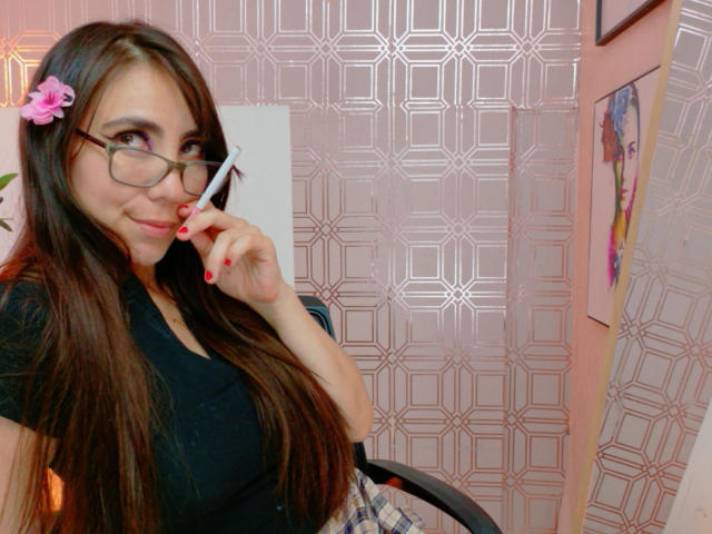 FreyaraVenwo with long brown hair, pink flower hairclip, glasses, black top, holding pen to mouth, red nail polish, pink wallpaper, plaid skirt, smiling, sexy, cute.