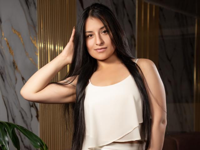 ElyThemsom heeft lang zwart haar, een bruine huid en kleine borsten, en draagt een witte crop top. Ze glimlacht, met haar hand in haar haar, en er staat een groene plant op de achtergrond..