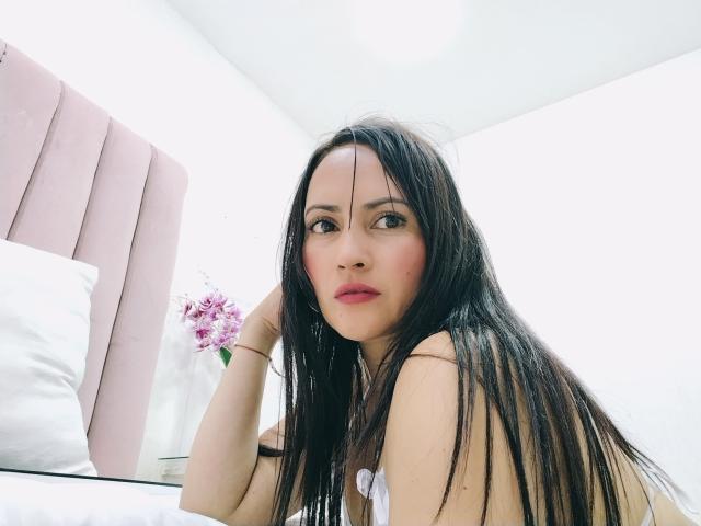 Emhily Diaz, con lunghi capelli neri, rossetto rosa e un'espressione seria, seduta su un letto con una testiera rosa, indossa una maglietta bianca e guarda la fotocamera..