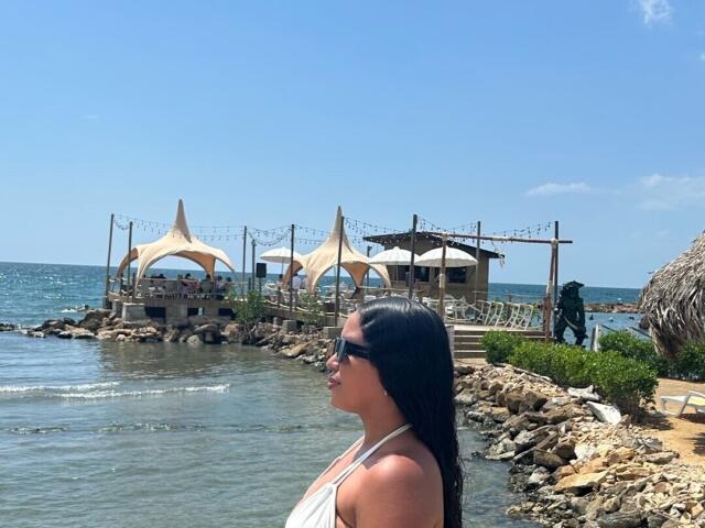 JeimyCampos has long black hair, wears sunglasses and a white halter top, standing by a rocky shoreline with a beach bar and umbrellas in the background.