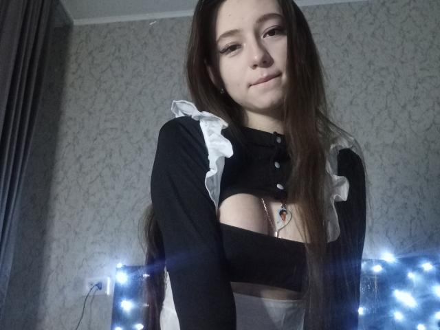 VeronicaAskwith with long brown hair, black and white maid outfit, small tits, smiling, wearing necklace, standing in front of blue string lights.