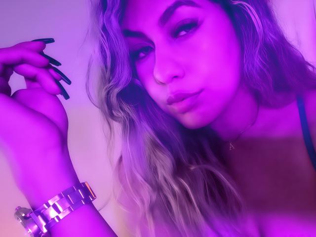 AnastaciaMonaco met paars aangelopen haar, lange zwarte nagels, een metalen armband en ketting, die in de camera kijkt met volle lippen, in een selfie met paars licht..