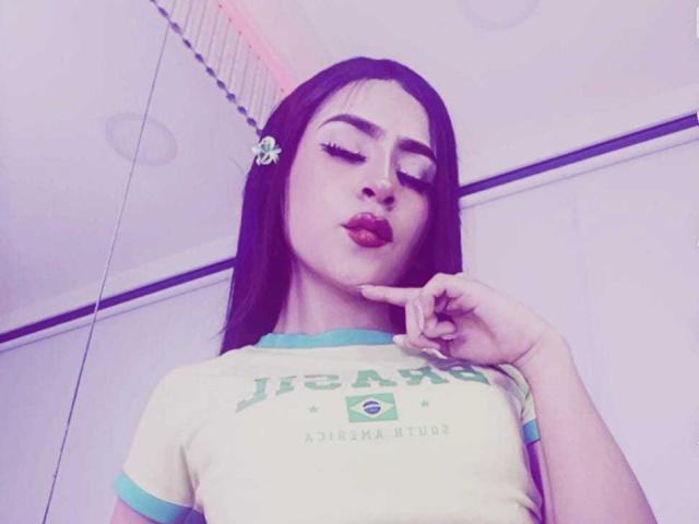 KimiNaru with long black hair, wearing a skull hair clip, pouting lips, and a yellow-green shirt, points to her lips with a finger. Her eyes are closed.