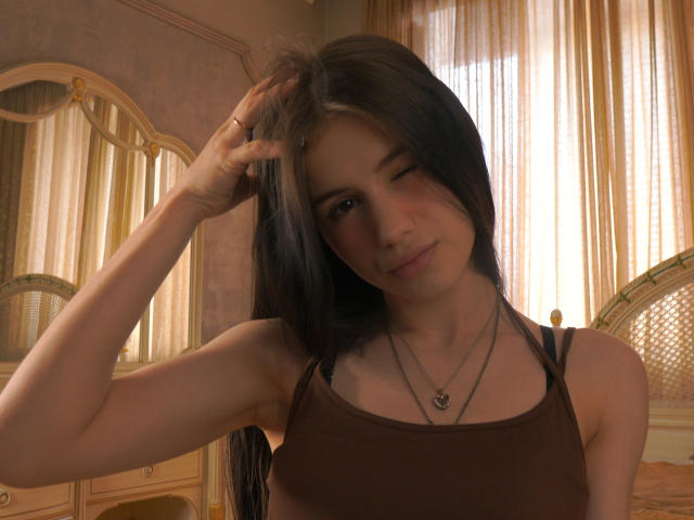 LatashaBasset with long black hair, winking, wearing a brown tank top, touching her head with one hand, showing small tits, and a necklace with a heart pendant.