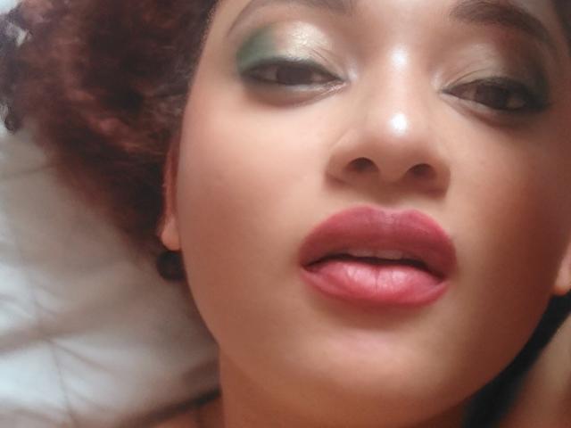 VioletaSwitch's face with green eyeshadow, slightly open pink lips, and curly brown hair, lying on a white pillow. Her nose is centered, and she's wearing black earbuds.