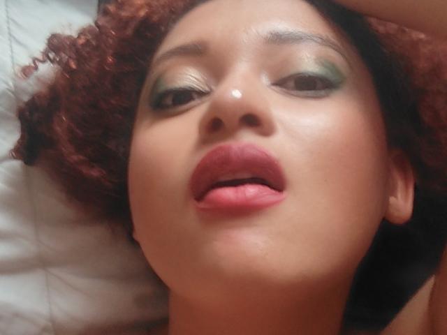 VioletaSwitch's face, lying down, with curly brown hair, green eyeshadow, and pink lips, tongue slightly out, looking up with a relaxed expression. Her skin is smooth and tan.