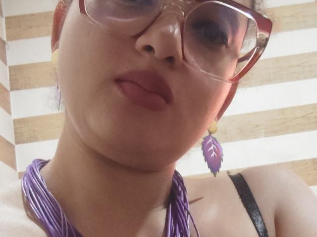 VioletaSwitch has a cute face, wearing purple glasses, purple feather earrings, and a purple necklace, with pink lips and a black strap top, showing a bit of her tits.