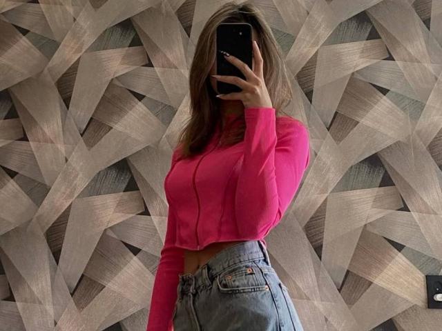LaunaGutterman usando uma cropped rosa de manga comprida, mostrando seios pequenos, vestindo jeans de cintura alta, segurando um celular, cobrindo o rosto, em pé em frente a uma parede estampada..
