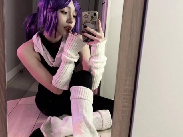 PaulineBitah, con cabello morado, vestida de negro, con un suéter blanco sin mangas, calentadores blancos en las piernas, pantalones negros, sentada en el suelo, tomando una selfie con el espejo, sosteniendo el teléfono, mirando a la cámara, con expresión de labios fruncidos..