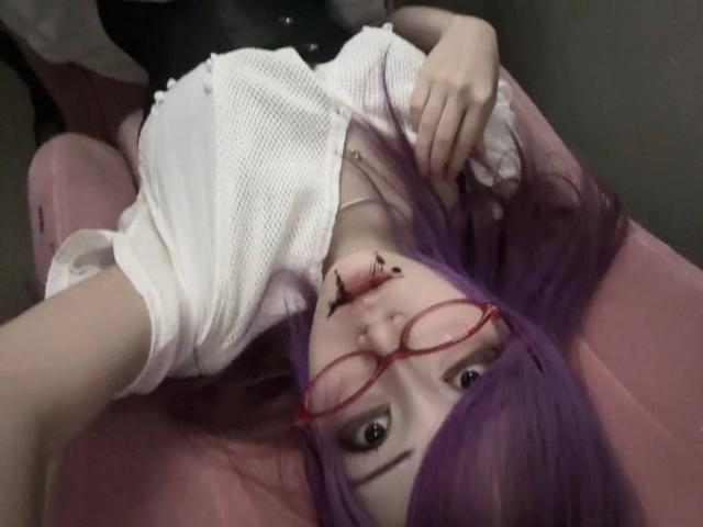 PaulineBitah, con i capelli viola, gli occhiali rossi e una camicia bianca, sdraiata su una sedia rosa, con del trucco nero sul naso, che tiene la camicia e guarda verso l'alto..