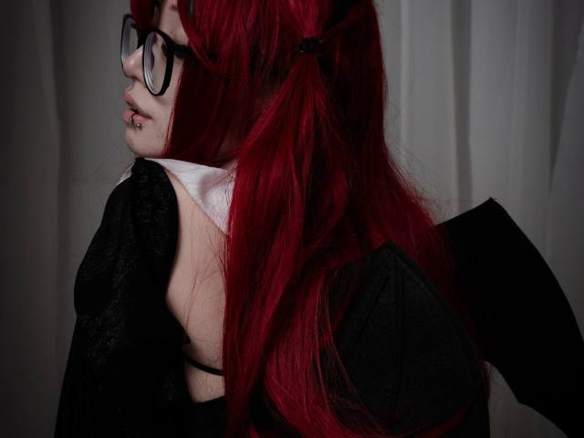 PaulineBitah, con i capelli rossi, gli occhiali neri e una maglia nera in pizzo, che mostra la sua schiena chiara e il colletto bianco, con un piercing al naso e alle labbra. Il suo seno è leggermente visibile..