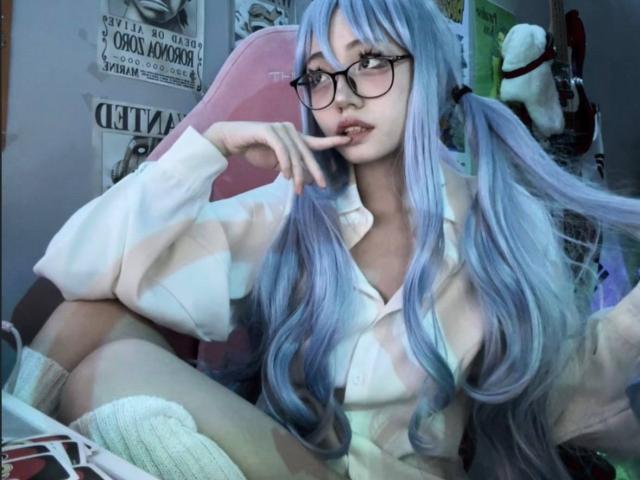 PaulineBitah, con i suoi lunghi capelli blu, gli occhiali, la camicia bianca e i calzini alti, è seduta con un dito sulle labbra, mostrando il seno. È seduta su una sedia rosa..