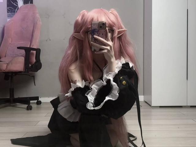 PaulineBitah with pink hair, elf ears, black and white frilled dress, holding phone, taking mirror selfie, kneeling, visible bare shoulder, pink chair, wooden floor, black cat toy.