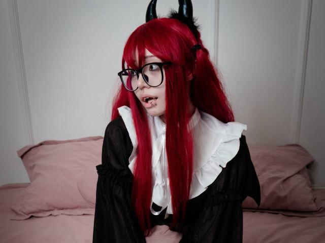 PaulineBitah met rood haar, zwarte hoorns, een bril en een zwarte jurk met witte ruches, die er verrast uitziet en kleine hoektanden laat zien, liggend op een roze bed..