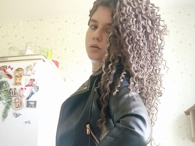 MarlanViviane, met krullend bruin haar, draagt een zwarte leren jas en staat in een keuken. Ze kijkt opzij, haar handen zitten in haar zakken. Op de achtergrond staat een witte koelkast met diverse decoraties..