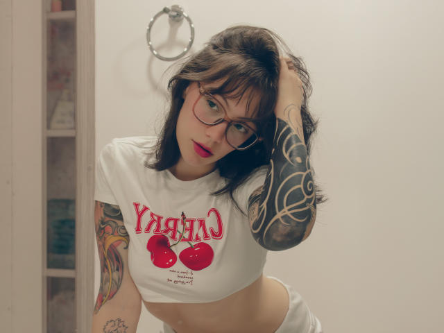 Isabella Arias con gli occhiali, top corto bianco con stampa di ciliegie rosse, tatuaggi sulle braccia, mano tra i capelli castani, in posizione inclinata in avanti, biancheria intima bianca, anello sul muro, rossetto rosso, che guarda verso la fotocamera..