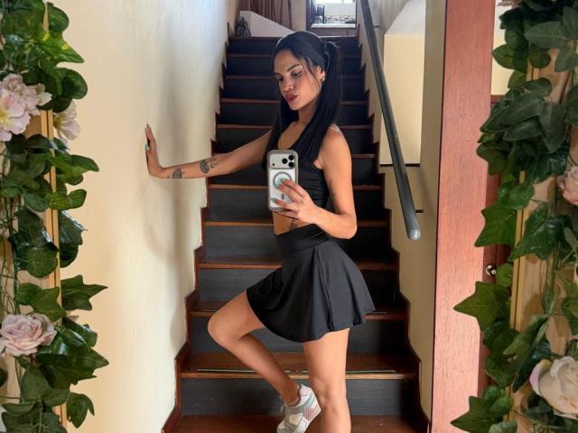 VicktoriaSunshine, avec des cheveux noirs, portant un haut court noir et une jupe, tenant un téléphone, debout sur des marches en bois, avec un tatouage sur le bras gauche, des chaussettes blanches, et des fleurs de chaque côté..