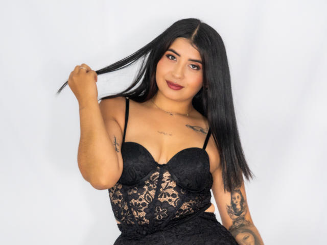 Valery Silva, con lunghi capelli neri, indossa un top di pizzo nero, tiene una ciocca di capelli, mostra tatuaggi sulle braccia e sul petto, sorride con il rossetto rosso..