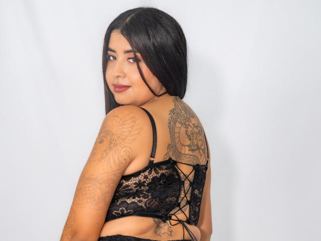 Valery Silva, con lencería de encaje negro, mirando por encima del hombro, mostrando brazos y espalda tatuados, con cabello negro largo y una ligera sonrisa, resaltando sus pechos y glúteos..