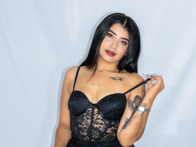 ValerySilva with long black hair, wearing a black lace bra, pulling a strap, showing tattoos on arms and chest, red lipstick, and a confident expression.