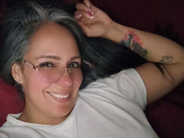 LailaCastillo, avec des cheveux gris, des lunettes, un t-shirt blanc, souriante, avec un tatouage sur le bras, sur un fond rouge, allongée, une main dans les cheveux, mignonne, sexy, attirante..