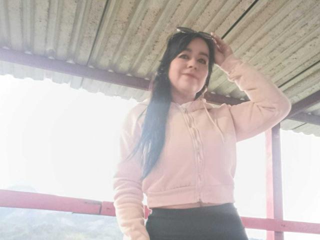 TamaraFoxy with long black hair, wearing a pink hoodie and black pants, standing under a corrugated metal roof, hand in hair, smiling slightly. Her ass is not visible.
