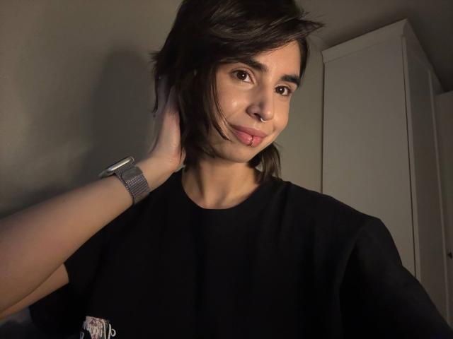 TameikaMacclellan with short dark hair, cute nose ring, black shirt, smirking, touching head with hand, silver watch, light skin, subtle makeup, in dimly lit room.